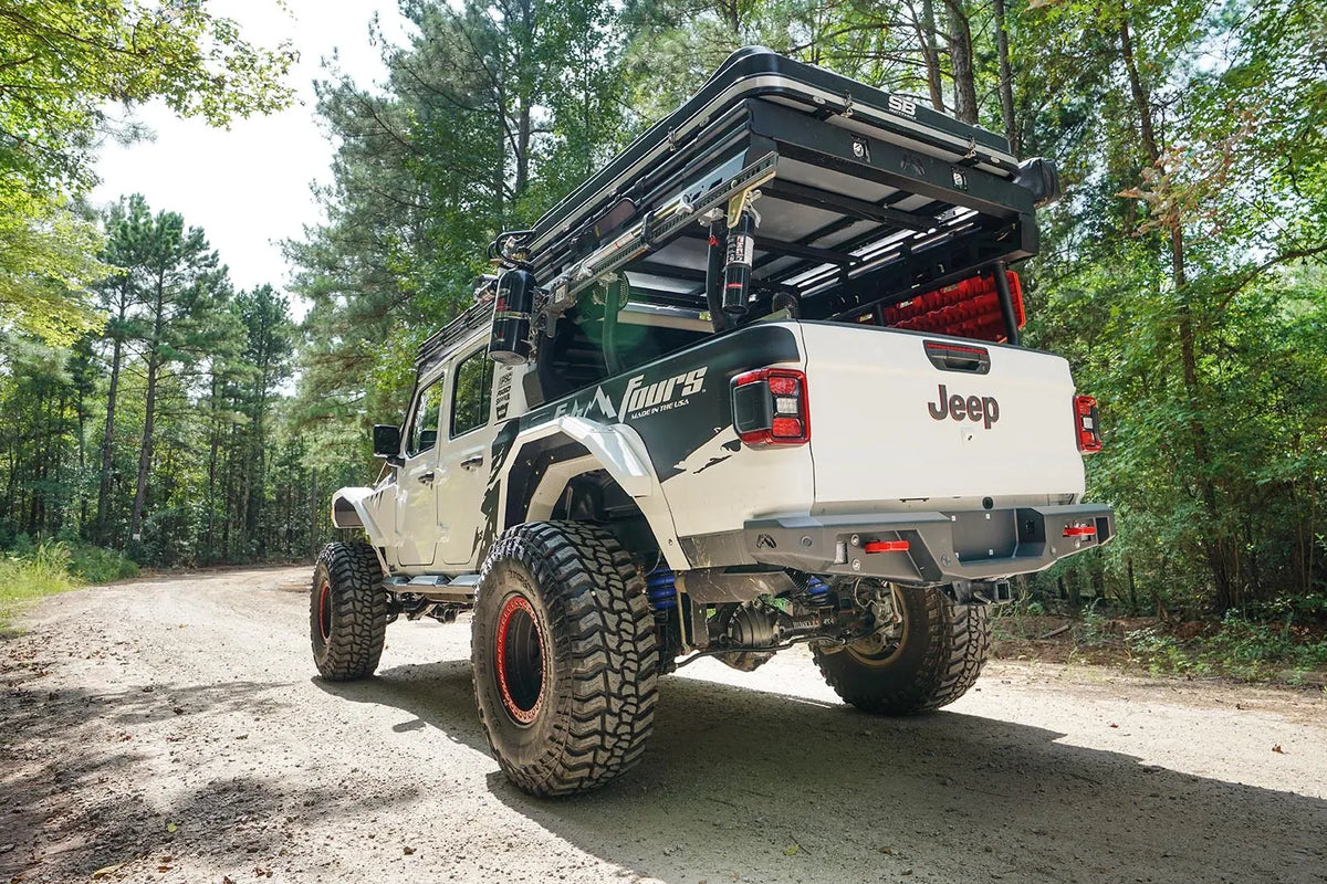 Fab Fours standard rear bumper on a lifted white Jeep Gladiator with Fab Fours graphics and overland bed rack, driver-side rear three-quarter view on a gravel road in the woods