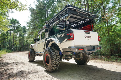 Fab Fours standard rear bumper on a lifted white Jeep Gladiator with Fab Fours graphics and overland bed rack, driver-side rear three-quarter view on a gravel road in the woods