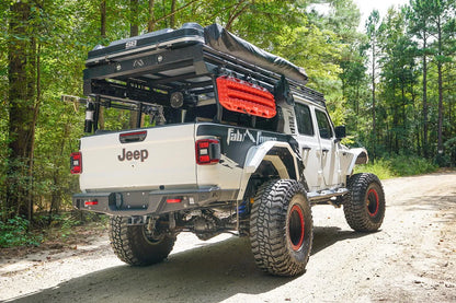 Fab Fours standard rear bumper on a lifted white Jeep Gladiator with Fab Fours graphics and overland bed rack, passenger-side rear three-quarter view on a gravel road in the woods