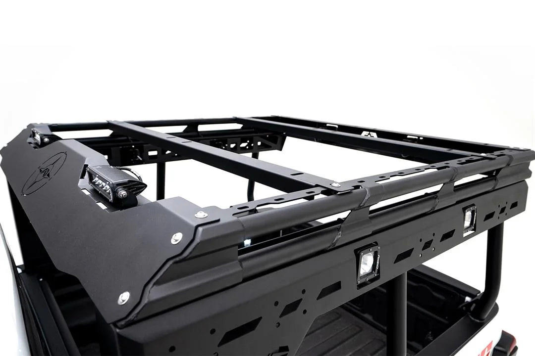 Close-up of the Fab Fours Overland Bed Rack on a Jeep Gladiator, showing the front fairing with compass logo and LED light, two cross-members and slotted side rail with RIGID cube lights