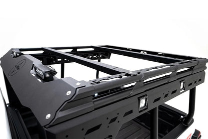 Close-up of the Fab Fours Overland Bed Rack on a Jeep Gladiator, showing the front fairing with compass logo and LED light, two cross-members and slotted side rail with RIGID cube lights
