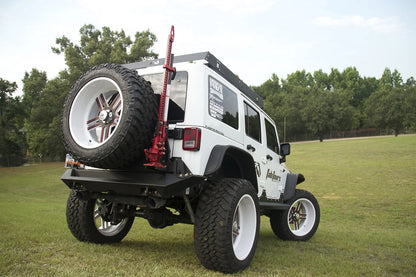 Fab Fours Hi-Lift jack mount on a white Jeep Wrangler JK on grass, rear passenger-side three-quarter view, showing the jack on the tire carrier, spare tire and rear bumper
