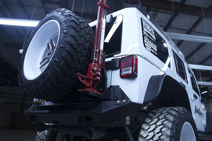 Fab Fours Hi-Lift jack mount on a white Jeep Wrangler JK, low rear passenger-side three-quarter view, showing the jack mounted to the off-the-door tire carrier beside the spare tire
