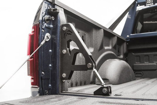 Fab Fours truck bed brace installed at the rear corner of a dark blue pickup bed, showing the matte black steel A-frame bracket with Fab Fours logo cutout and adjustable turnbuckle link bolted to the bed floor