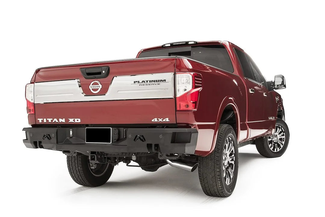 Fab Fours Premium rear bumper on a red Nissan Titan XD, passenger-side rear three-quarter view, with parking sensors, Fab Fours logo cutouts, and hitch receiver