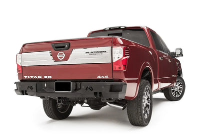 Fab Fours Premium rear bumper on a red Nissan Titan XD, passenger-side rear three-quarter view, with parking sensors, Fab Fours logo cutouts, and hitch receiver