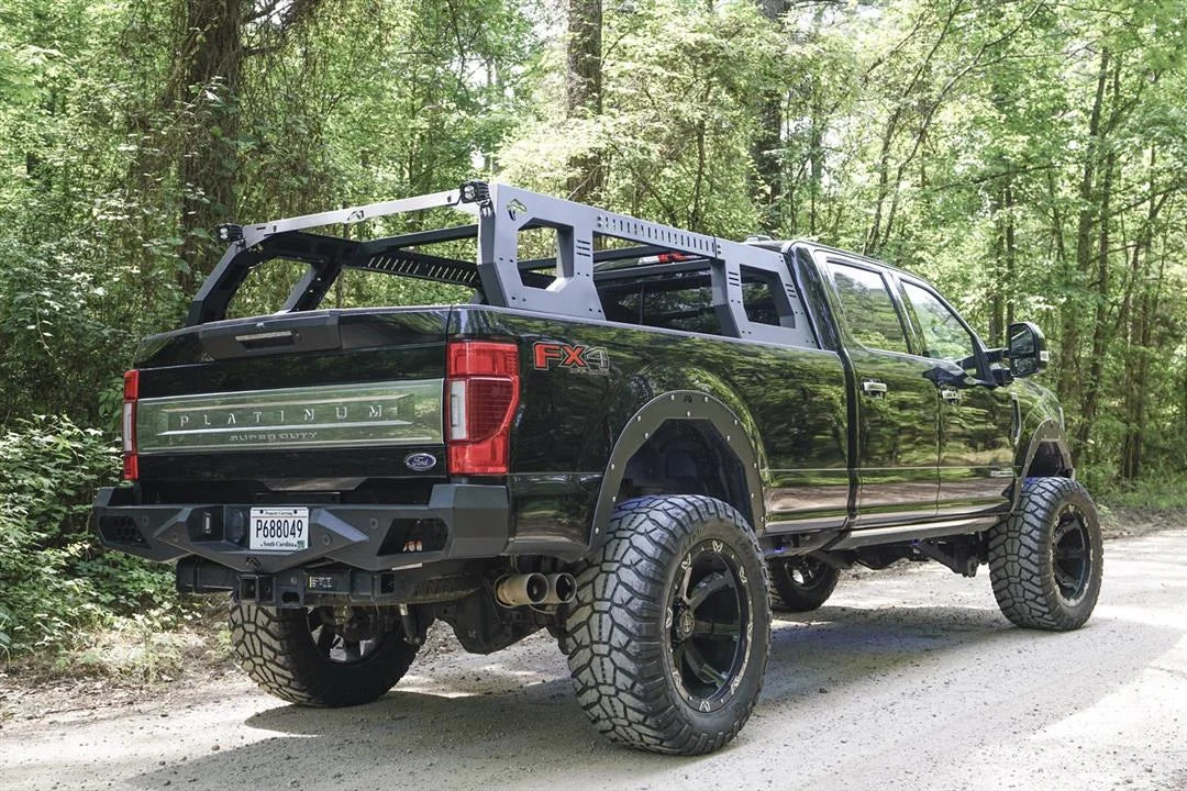 Fab Fours Adjustable Overland Rack on a black full-size pickup truck on a forest road, rear passenger-side view, showing the matte black rack with cube lights over the bed