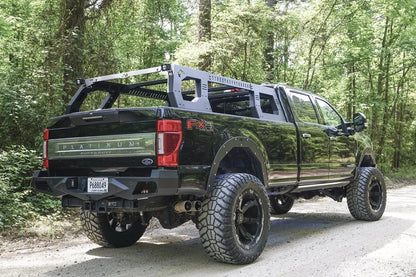 Fab Fours Adjustable Overland Rack on a black full-size pickup truck on a forest road, rear passenger-side view, showing the matte black rack with cube lights over the bed