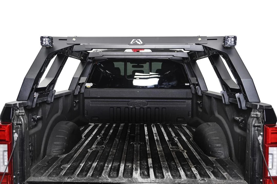 Fab Fours Adjustable Overland Rack in a full-size pickup bed, rear view through the open tailgate, showing the logo crossbar with two cube lights and the angled side frames