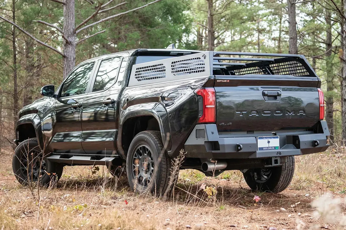 Fab Fours half rack with perforated side panels on a black Toyota Tacoma in the woods, driver-side rear three-quarter view, with Fab Fours rear bumper