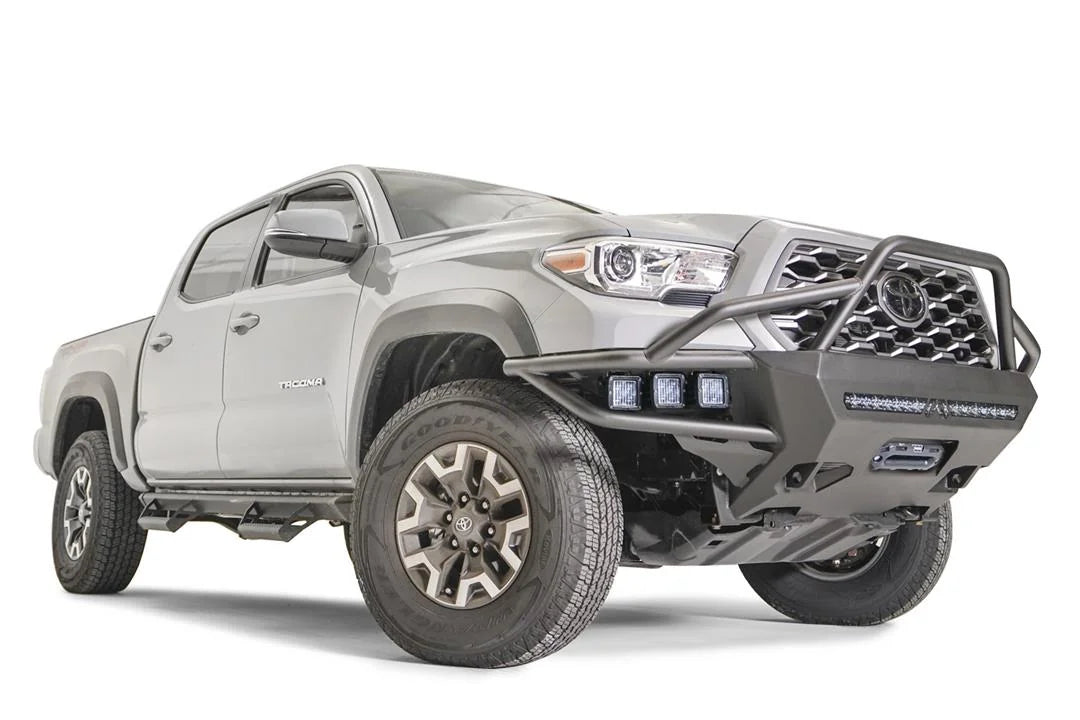 Fab Fours Hybrid front bumper with full guard on a silver Toyota Tacoma, front three-quarter view from the passenger side, showing the full grille guard, integrated pod lights, LED light bar and skid plate