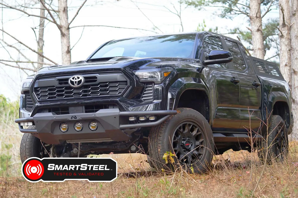 Fab Fours Ultra Light front bumper on a black Toyota Tacoma, front three-quarter view from the passenger side outdoors, showing the tube wings, three round lights and LED pod lights