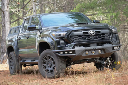 Fab Fours Ultra Light front bumper on a black Toyota Tacoma, front three-quarter view from the passenger side outdoors, showing the tube wings, three round lights and LED pod lights
