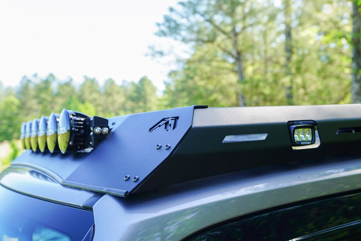 Close-up of the Fab Fours Roof Rack wind fairing on a Toyota Land Cruiser, showing the Fab Fours logo cutout, RIGID round lights and a side-mounted RIGID cube light