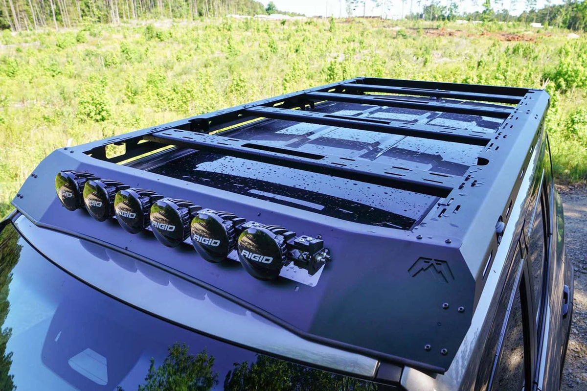 Fab Fours Roof Rack on a gray Toyota Land Cruiser outdoors, elevated driver-side view, showing the platform crossbars and six RIGID lights across the logo-cut wind fairing