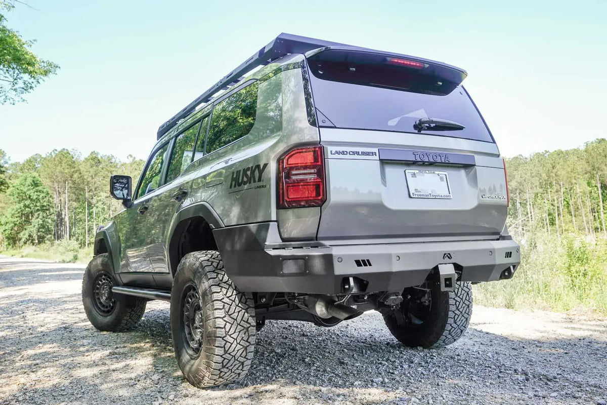 Fab Fours Premium rear bumper on a lifted silver Toyota Land Cruiser with roof rack, driver-side rear three-quarter view on a gravel road, with hitch receiver