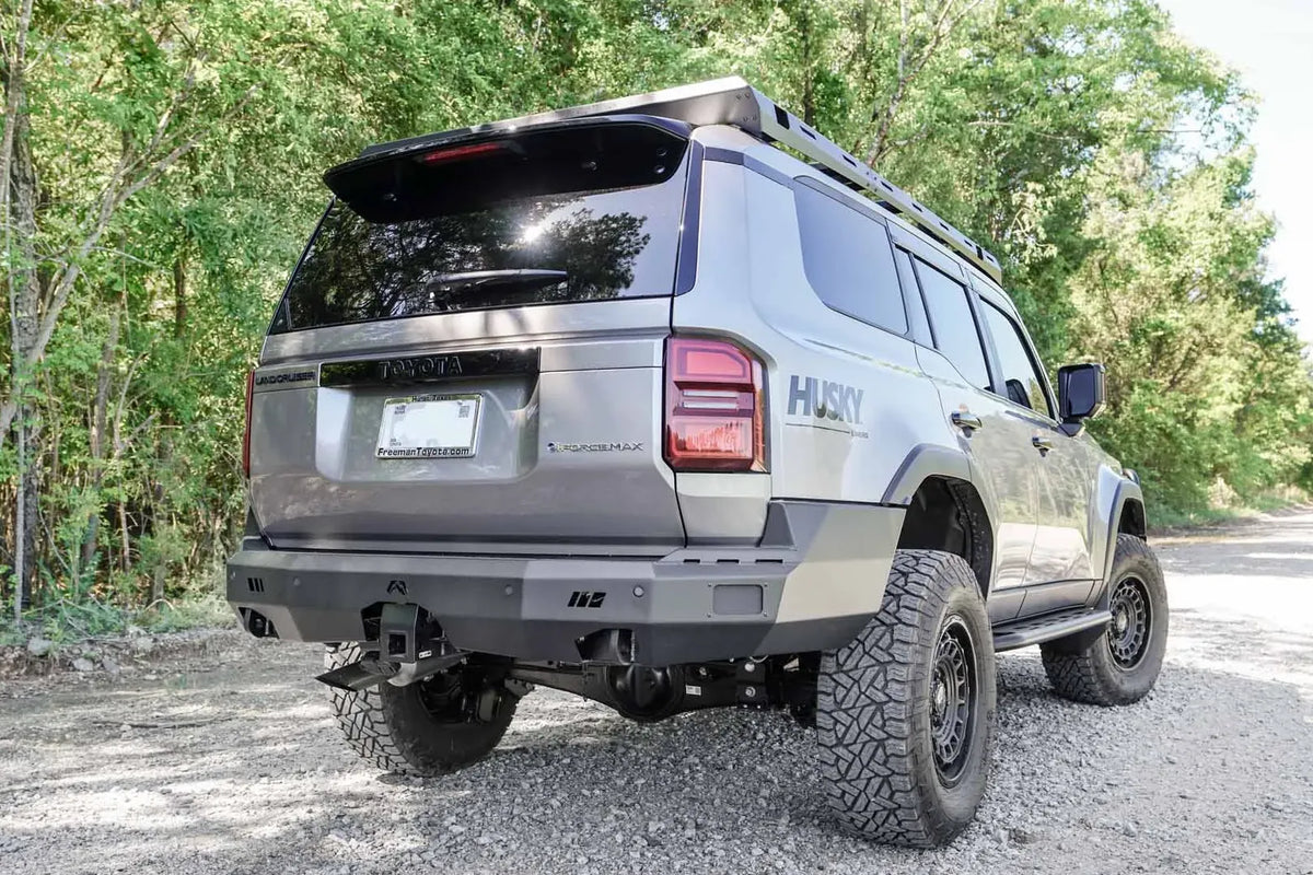 Fab Fours Premium rear bumper on a lifted silver Toyota Land Cruiser with roof rack, passenger-side rear three-quarter view on a gravel road, with hitch receiver
