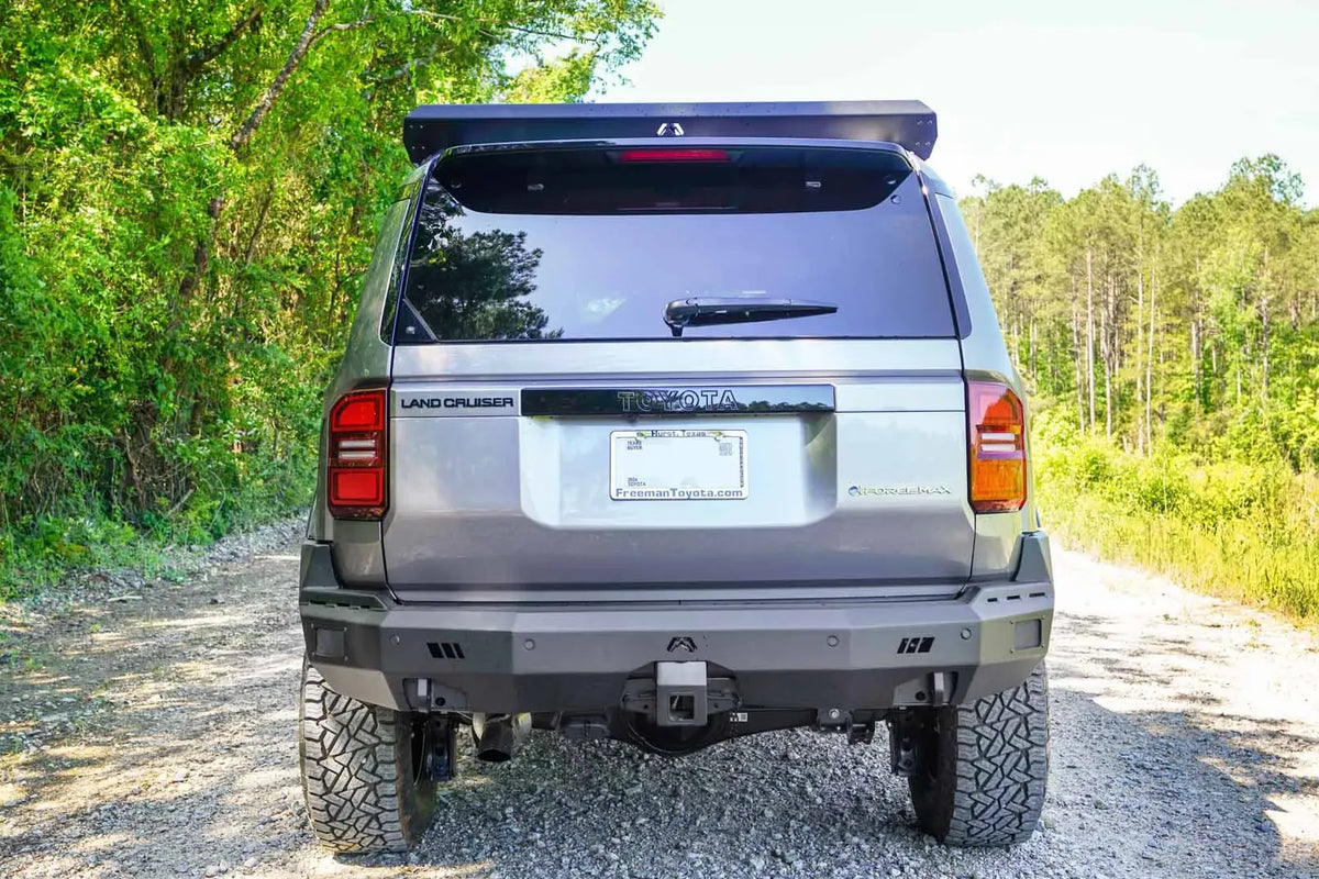 Fab Fours Premium rear bumper on a silver Toyota Land Cruiser on a gravel road in the woods, straight rear view, with parking sensors and hitch receiver