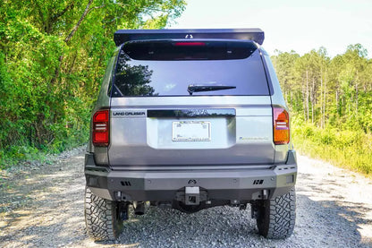 Fab Fours Premium rear bumper on a silver Toyota Land Cruiser on a gravel road in the woods, straight rear view, with parking sensors and hitch receiver