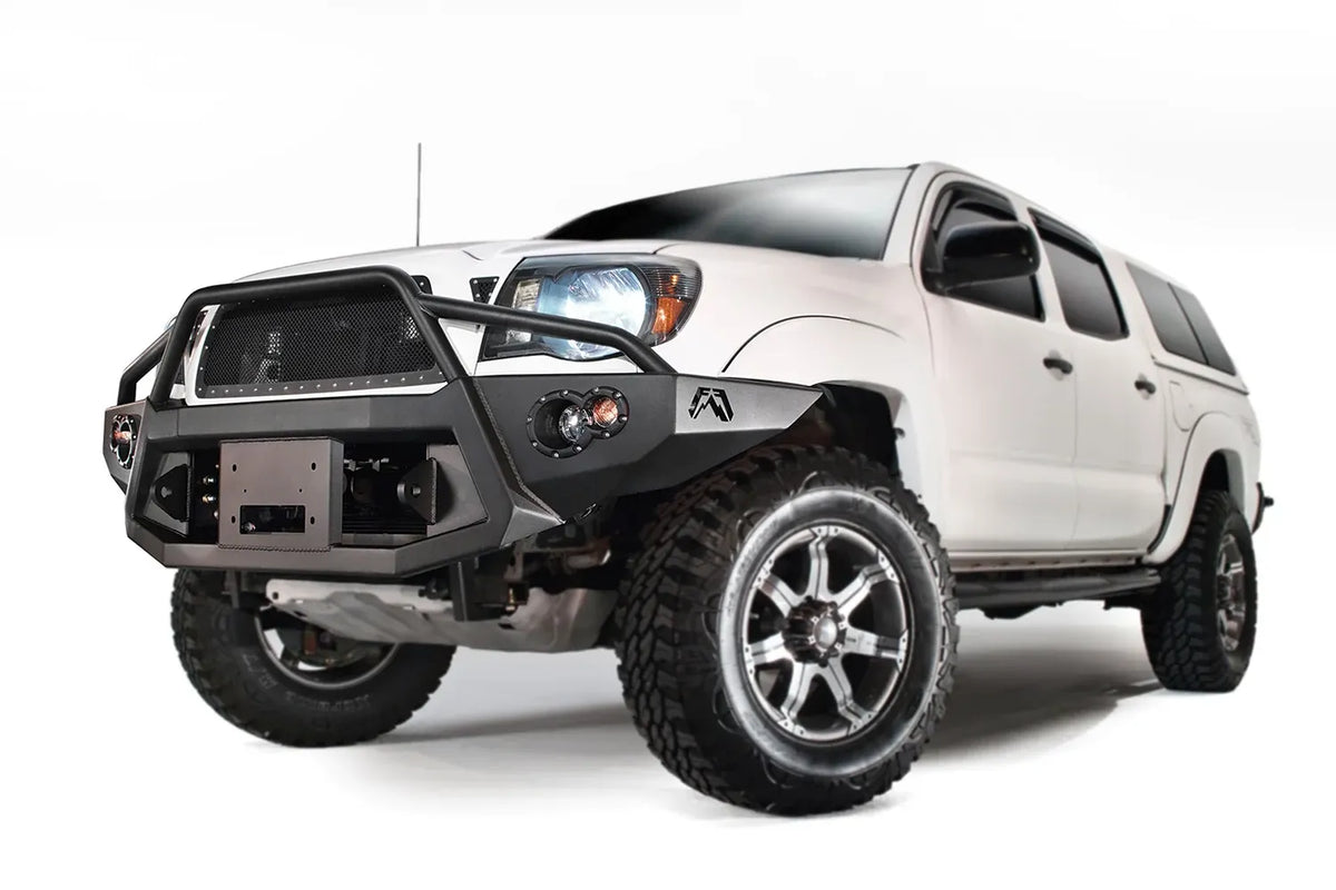 Fab Fours Premium front bumper with pre-runner guard on a white Toyota Tacoma, front three-quarter view from the driver side, showing the pre-runner hoop, recessed winch plate and round fog lights