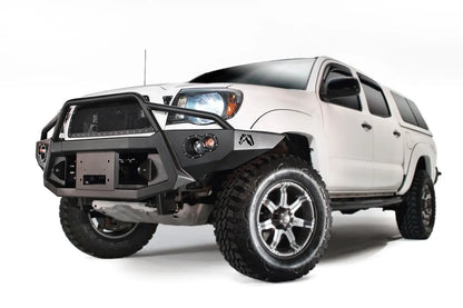 Fab Fours Premium front bumper with pre-runner guard on a white Toyota Tacoma, front three-quarter view from the driver side, showing the pre-runner hoop, recessed winch plate and round fog lights