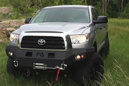 Fab Fours Premium front bumper on a silver Toyota Tundra, front view outdoors, showing the winch with red strap, D-ring shackles and round fog lights
