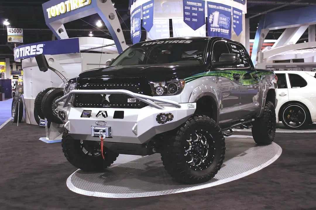 Fab Fours Premium front bumper with pre-runner guard on a black and silver Toyota Tundra, front three-quarter view from the driver side at a show display, showing the pre-runner hoop, winch and round fog lights