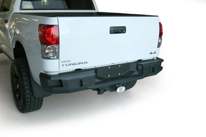 Fab Fours Premium rear bumper on a white Toyota Tundra, rear three-quarter view from the driver side in a garage, showing the full-width bumper face, wrap-around end caps and receiver hitch
