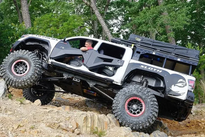 Fab Fours trail doors on a lifted white Toyota Tacoma climbing a rocky trail, driver-side view, showing the faceted front and rear doors with the driver's arm resting on the open top
