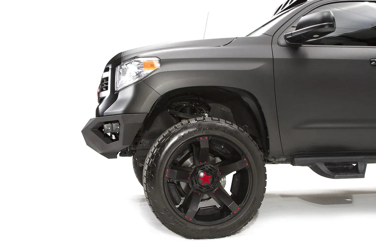 Fab Fours Vengeance front bumper on a matte black Toyota Tundra, close-up of the front corner from the driver side, showing the angled bumper end and wheel clearance