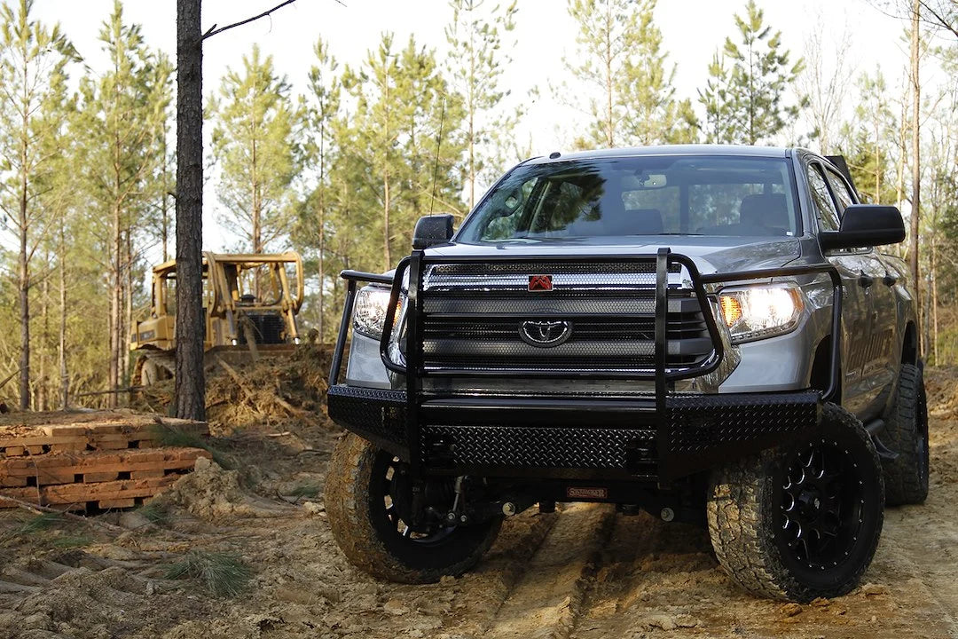 Fab Fours Black Steel front bumper with full guard on a gray Toyota Tundra, front view outdoors in a wooded clearing, showing the full grille guard, diamond-plate face, and side wings
