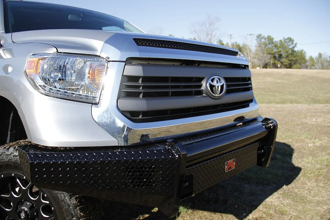 Fab Fours Black Steel front bumper on a silver Toyota Tundra, close-up corner view outdoors, showing the diamond-plate face, tubular top bar, and logo badge