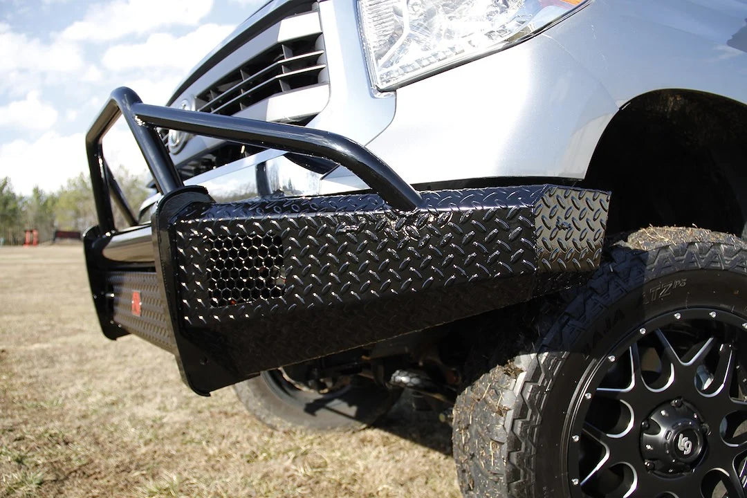 Fab Fours Black Steel front bumper with pre-runner guard on a silver Toyota Tundra, close-up corner view outdoors, showing the diamond-plate end cap, pre-runner tubing, and logo badge