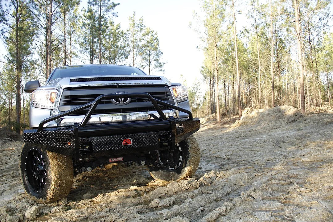 Fab Fours Black Steel front bumper with pre-runner guard on a silver Toyota Tundra, front view outdoors on a dirt trail, showing the pre-runner hoop, diamond-plate face, and side wings