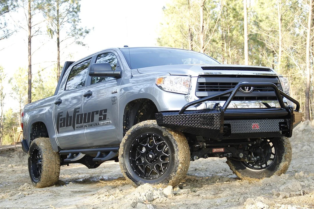 Fab Fours Black Steel front bumper with pre-runner guard on a silver Toyota Tundra, front three-quarter view outdoors on a dirt trail, showing the pre-runner hoop, diamond-plate face, and wraparound end caps