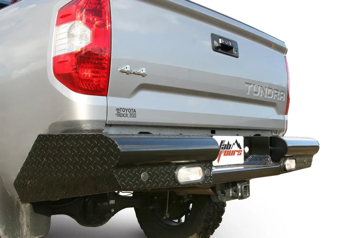 Fab Fours Black Steel rear bumper on a silver Toyota Tundra, rear three-quarter view outdoors, showing the diamond-plate face, recessed reverse lights, and wraparound end cap