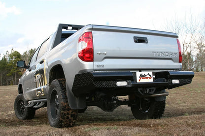Fab Fours Black Steel rear bumper on a silver Toyota Tundra, rear three-quarter view outdoors, showing the diamond-plate face, recessed reverse lights, and license plate mount
