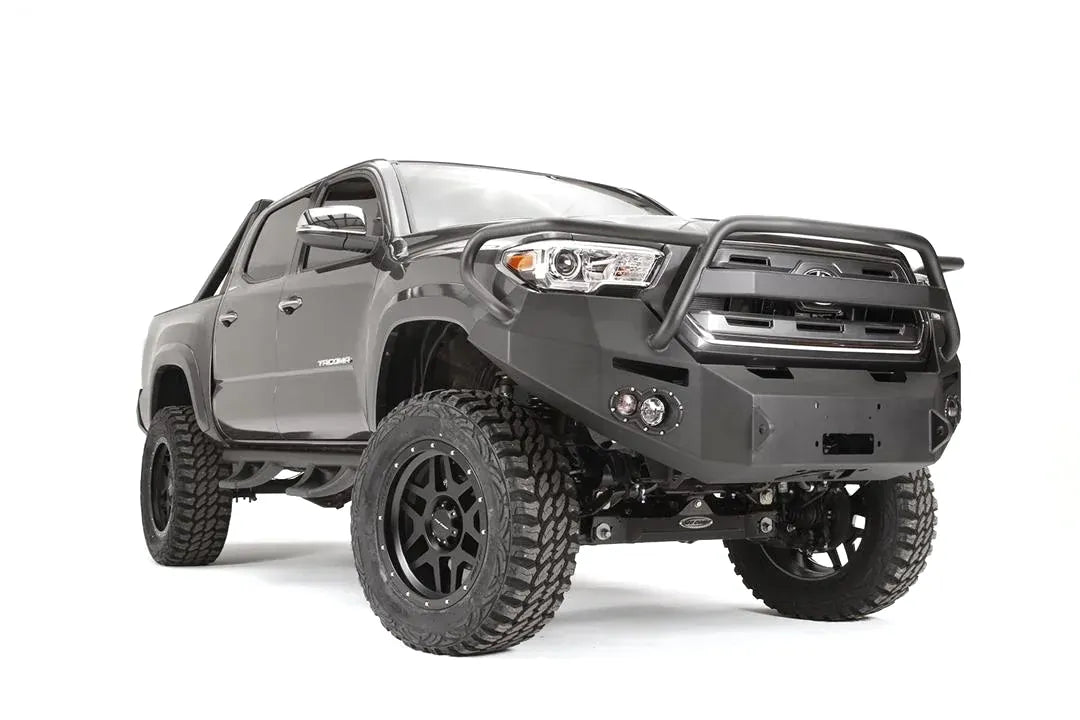 Fab Fours Premium front bumper with pre-runner guard on a dark gray Toyota Tacoma, front three-quarter view, showing the guard hoop, round fog lights, and winch-ready center section