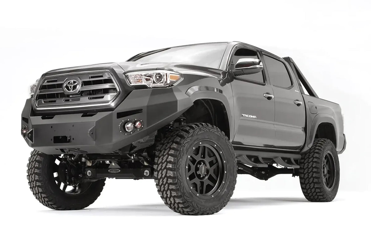 Fab Fours Premium front bumper on a dark gray Toyota Tacoma, front three-quarter view, showing the winch-ready center section, round fog lights, and wraparound end caps