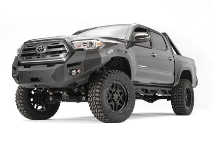 Fab Fours Premium front bumper on a dark gray Toyota Tacoma, front three-quarter view, showing the winch-ready center section, round fog lights, and wraparound end caps