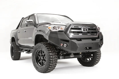 Fab Fours Premium front bumper on a dark gray Toyota Tacoma, front three-quarter view, showing the no-guard design, round fog lights, and winch mount plate