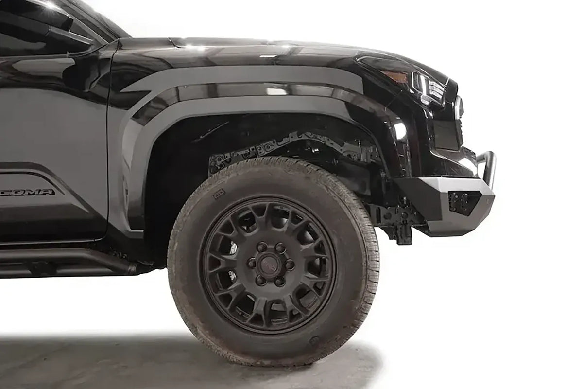 Fab Fours Vengeance front bumper with pre-runner guard on a black Toyota Tacoma, close-up side profile of the front corner, showing the bumper end cap, guard tubing, and wheel clearance