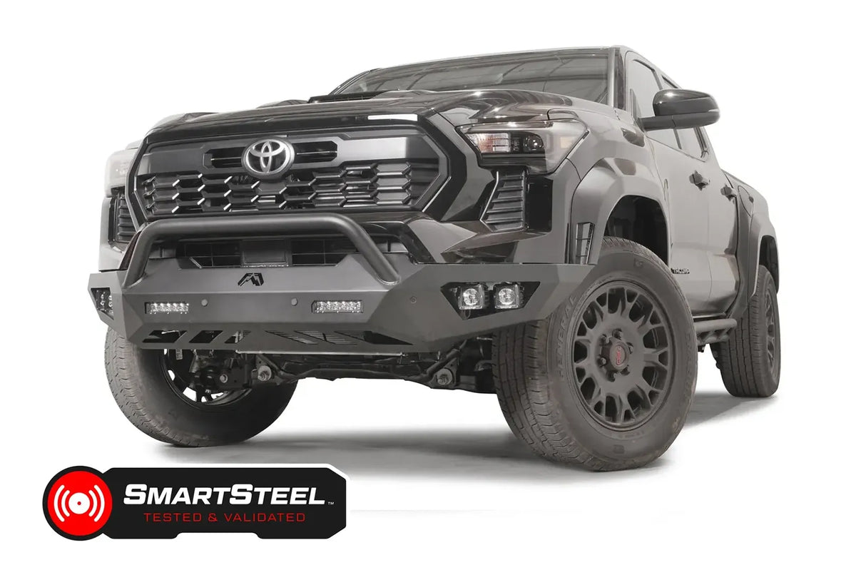 Fab Fours Vengeance front bumper with pre-runner guard on a dark gray Toyota Tacoma, front three-quarter view, showing the pre-runner hoop, LED light bars, and cube light pods
