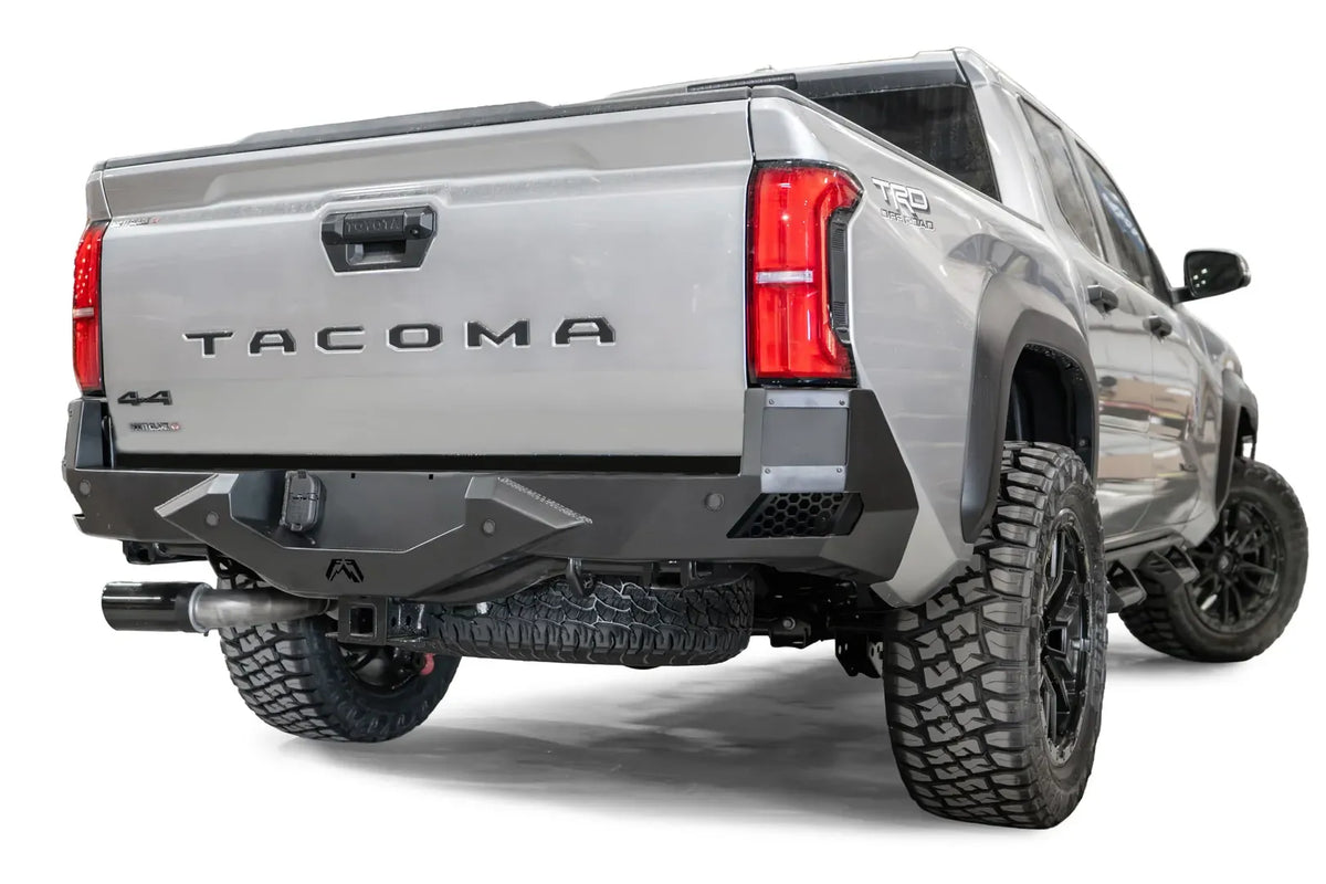 Fab Fours Vengeance rear bumper on a silver Toyota Tacoma, rear three-quarter view, showing the angular faceted face, recessed light pods, and receiver hitch