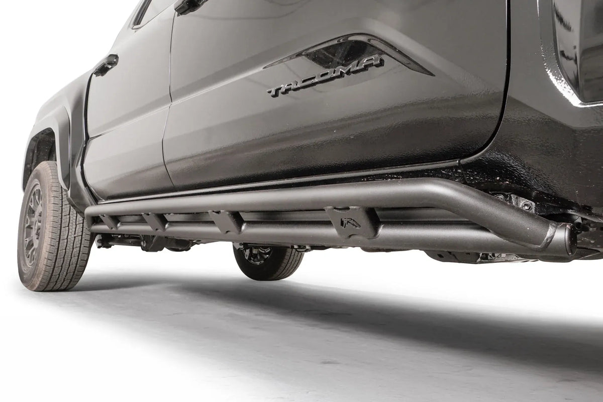 Fab Fours tube rock slider on a black Toyota Tacoma, low driver-side view, showing the matte black double-tube design with welded gussets and Fab Fours logo plate under the doors