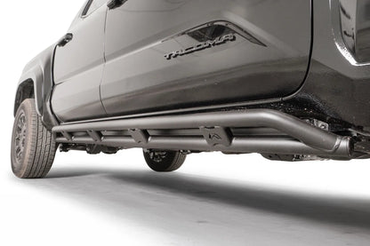Fab Fours tube rock slider on a black Toyota Tacoma, low driver-side view, showing the matte black double-tube design with welded gussets and Fab Fours logo plate under the doors