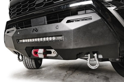 Fab Fours Hidden Winch Mount on a black Toyota Tacoma, close-up front view, showing the winch hook, LED light bar, and D-ring shackle