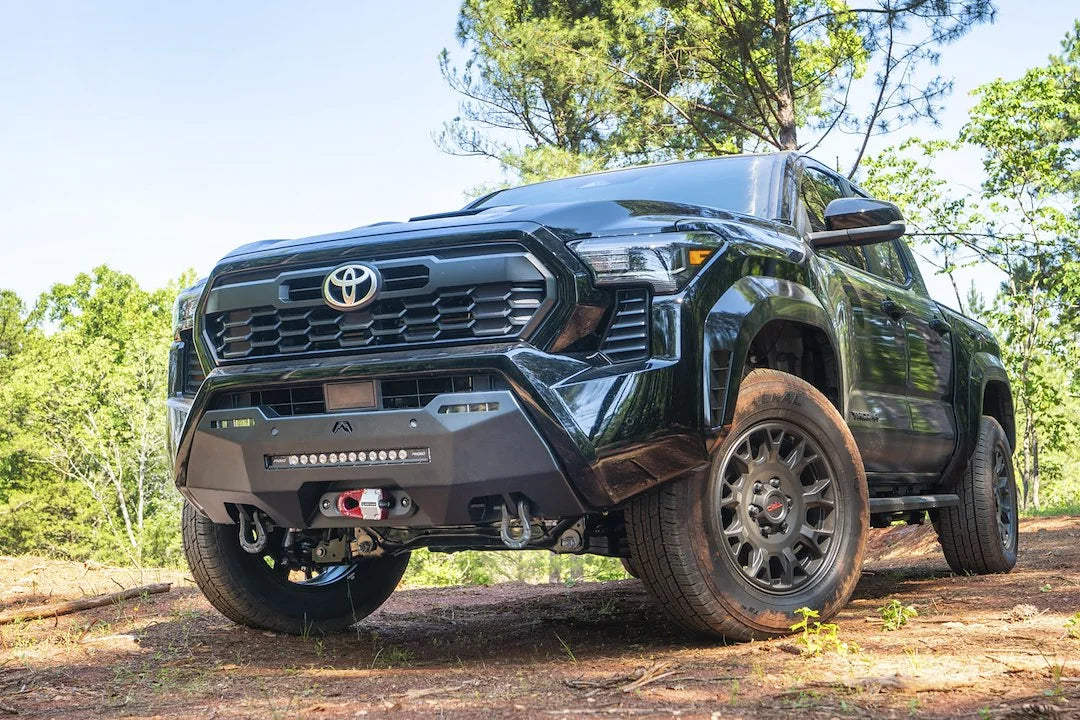 Fab Fours Hidden Winch Mount on a black Toyota Tacoma, front three-quarter view outdoors on a dirt trail, showing the winch fairlead, LED light bar, and D-ring shackles