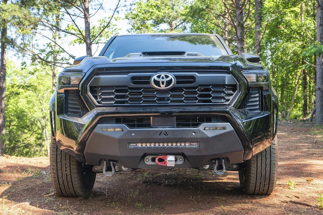 Fab Fours Hidden Winch Mount on a black Toyota Tacoma, front view outdoors in the woods, showing the winch with red hook, LED light bar, and D-ring shackles