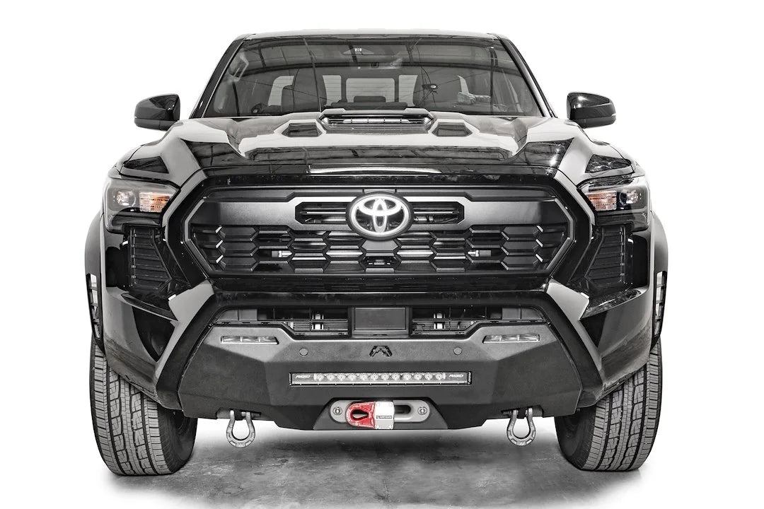 Fab Fours Hidden Winch Mount on a black Toyota Tacoma, straight-on front view, showing the hidden winch with red hook, LED light bar, and D-ring shackles